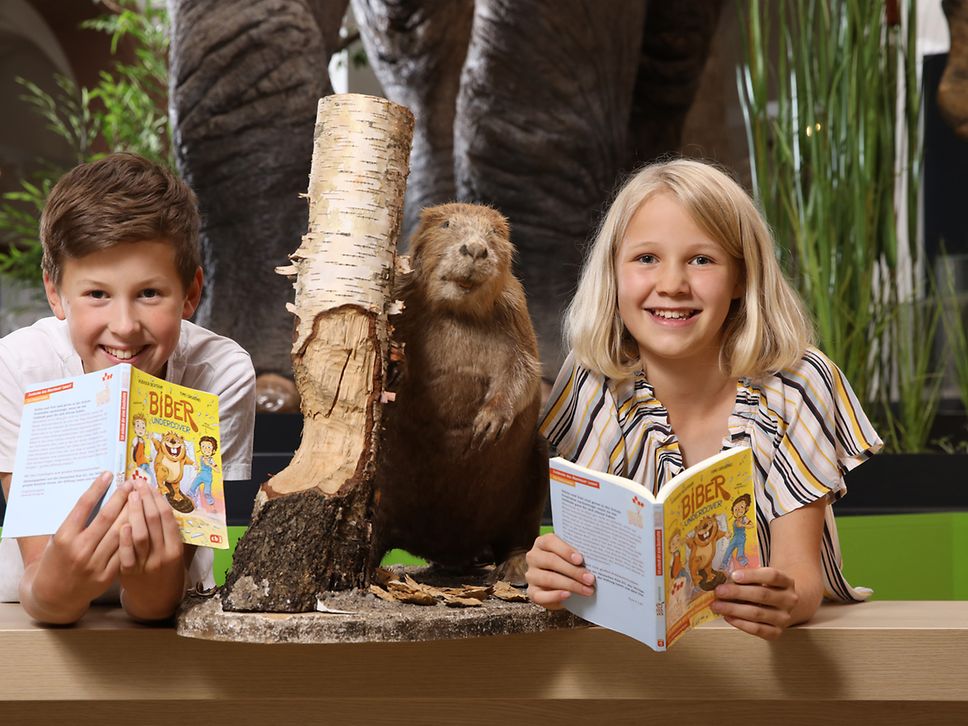 Zwei Kinder mit Büchern in der Hand, zwischen ihnen ist eine Biber-Figur