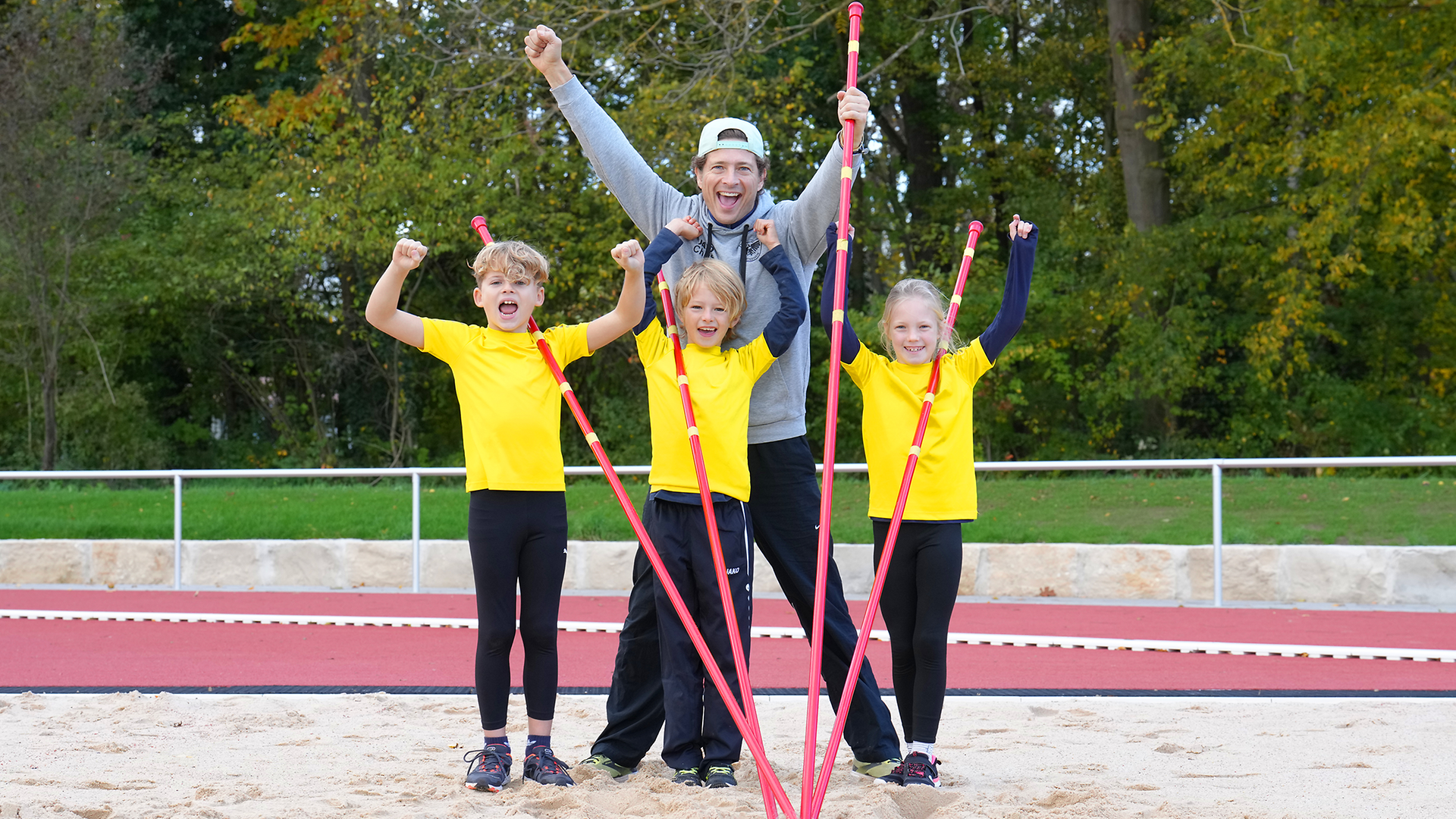 Ein Mann und drei Kinder stehen in einer Sandgrube auf einem Sportplatz und halten 4 Hochsprungstäbe in der Hand und jubeln